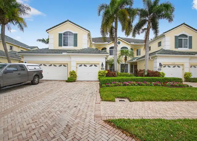 Yellow two-story townhomes with white garages, palm trees, and a brick driveway under blue skies.