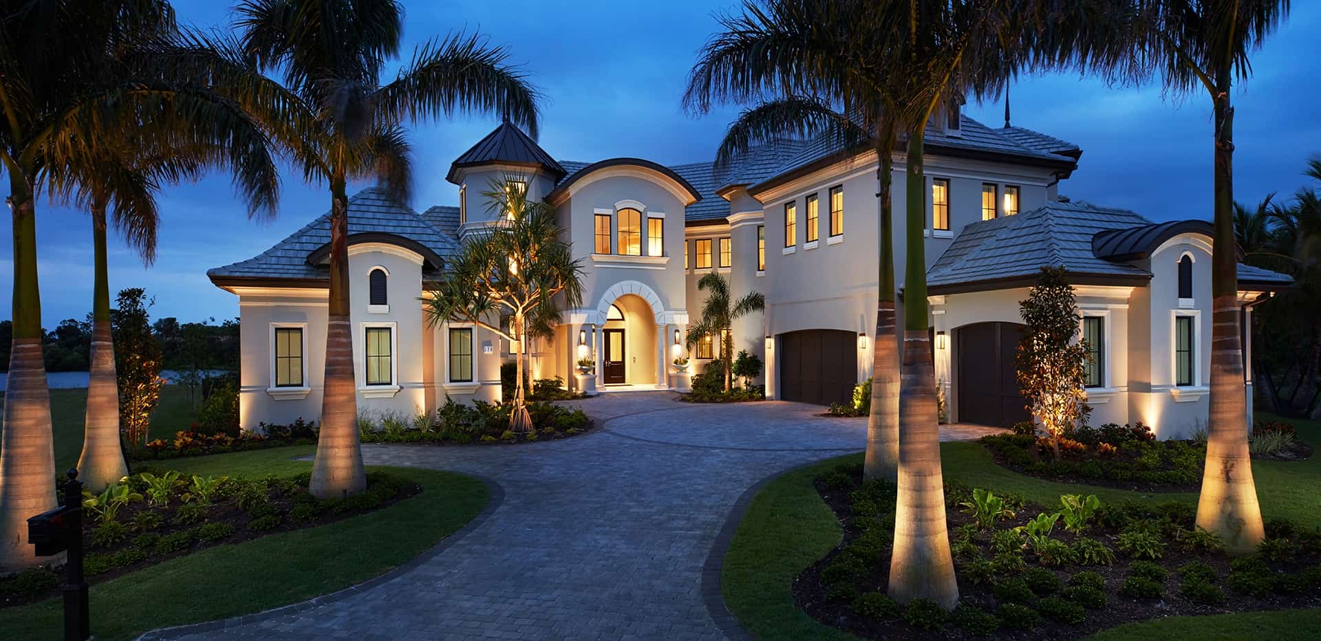 Illuminated luxury mansion at dusk featuring palm trees, warm lighting, and a circular stone driveway.