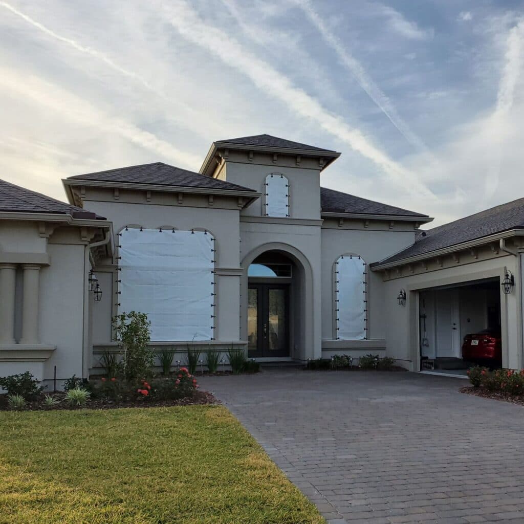 Beige house with white hurricane shutters on windows, brick driveway, and blue sky with contrails.